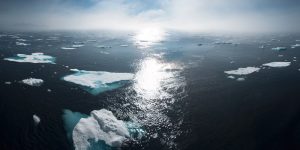 photo of ice bergs floating in the ocean. The sky is blue in the background and cloudy, and the sun is reflected in the middle of the image.